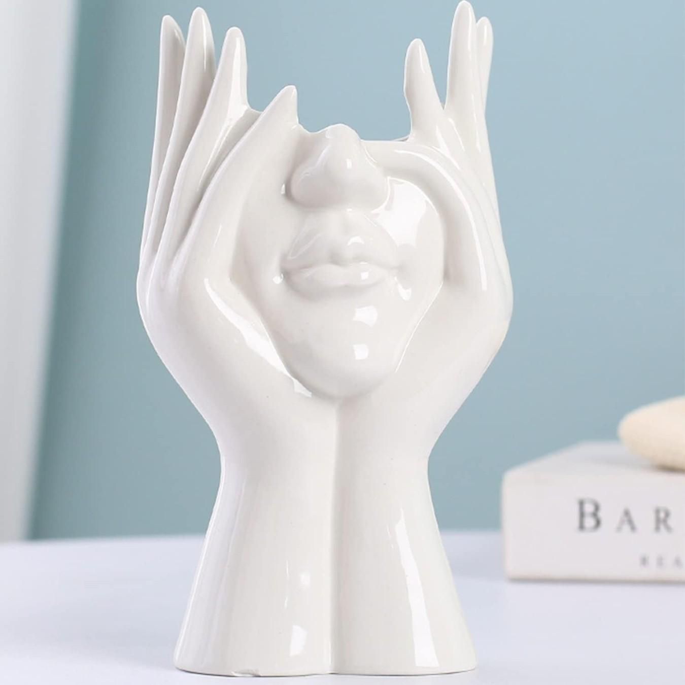White ceramic sculpture of hands holding a face against a light blue background