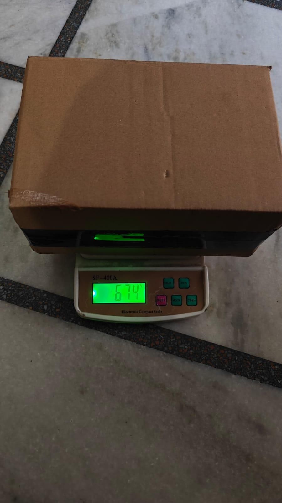 Cardboard box on a digital scale with a green display on a tiled floor.